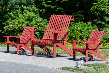 Three public red wooden chairs in the sunshine at a park in  Vancouver, British Columbia, Canada in the summer sun with green background.