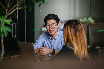 A pair of diverse team mates have a business meeting and discussion together to collaborate. A Korean man is sitting at a table in a trendy coworking space office and talking to his female companion.