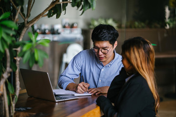 A pair of diverse team mates have a business meeting and discussion together to collaborate. A Korean man is sitting at a table in a trendy coworking space office and talking to his female companion.