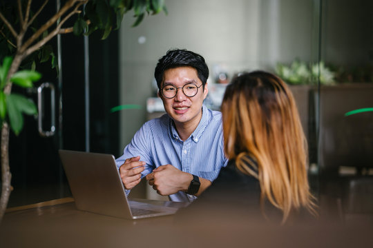 A pair of diverse team mates have a business meeting and discussion together to collaborate. A Korean man is sitting at a table in a trendy coworking space office and talking to his female companion.