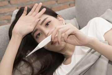 Naklejka premium Pretty Asian woman sick from cold and used with thermometer lying on sofa in the room.