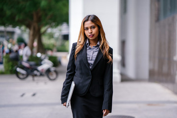 Fototapeta premium Portrait of a beautiful, young and attractive Singaporean Malay Asian woman in a business suit walking in a city in Asia during the day with her laptop computer. She is smiling confidently.