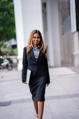 Portrait of a beautiful, young and attractive Southeast Asian woman in a business suit walking in a city in Asia during the day with her laptop computer. She is smiling confidently.