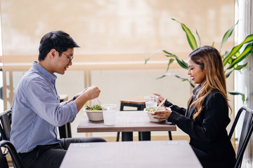 A pair of diverse colleagues sit and have a healthy salad lunch together. They are professionally dressed and chatting as they dig into their food. One is a Malay Asian woman, the other a Korean man.