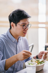 A Korean Asian man enjoys his salad lunch as he sits in a cafe in the city during the day. He is tall, handsome and is wearing a collared shirt and spectacles.