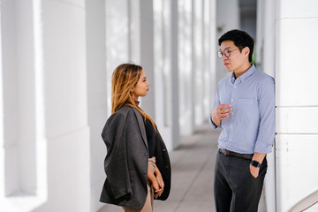 Candid shot of two diverse Asian business enthusiast, Korean man and a Malay woman having an animated conversation as they standby the hall during the day. They're both wearing a corporate attire.