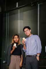 Business portrait of two diverse Asian business people (colleague meeting for a snack) standing by the cafe. One is a Korean man,the other a Malay woman. They're both smiling and holding their treats.