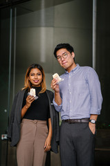 Business portrait of two diverse Asian business people (colleague meeting for a snack) standing by the cafe. One is a Korean man,the other a Malay woman. They're both smiling and holding their treats.