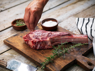 Hands curing a raw fresh angus meat tomahawk on a wooden cutting board with rosemary, salt and butcher's knife