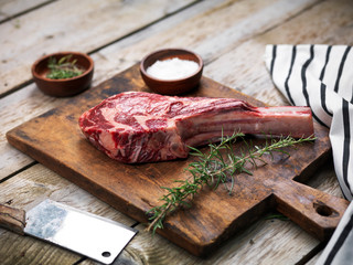 Raw fresh angus meat tomahawk on a wooden cutting board with rosemary, salt and butcher's knife