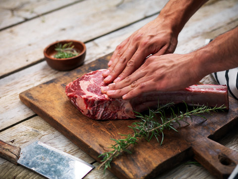 Hands curing a raw fresh angus meat tomahawk on a wooden cutting board with rosemary, salt and butcher's knife