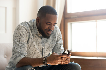 Young african american man using smartphone app sit on sofa