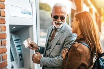 Senior father and his daughter using atm machine together to withdraw money. They are happy. Bright...