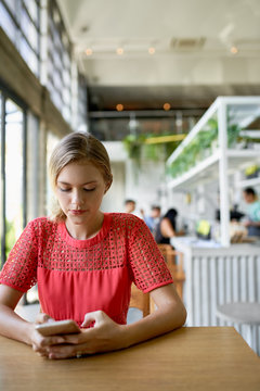 Lifestyle shot of young blonde caucasian natural millennial woman looking at cellphone at a modern tropical and bright restaurant with plants
