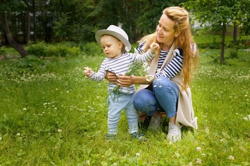 Fototapeta premium Happy mother with her little son in a big hat are walking on a green meadow in a summer park with a bouquet of chamomiles