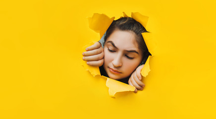 Funny angry teenage girl peeping through hole on yellow paper. The concept of surprise, skeptic, sad mood from what he saw. Discounts, sales, surprise. Copy space.