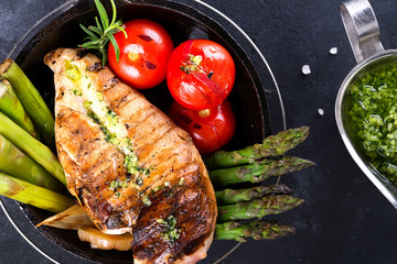 Grilled chicken breast on a cast iron skillet with grill vegetables and green sauce on a stone background, flat lay