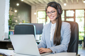 Smiling female customer support operator talking with client and using laptop in office