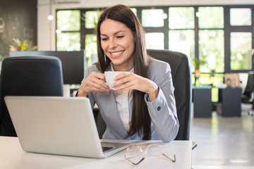 Smiling young businesswoman drinking coffee while using laptop in office