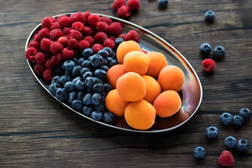 Fresh apricots, raspberries and blueberries on an oval metal plate. Gray background. Soft focus.