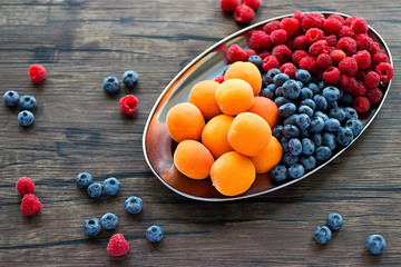 Fresh apricots, raspberries and blueberries on an oval metal plate. Gray background. Soft focus.