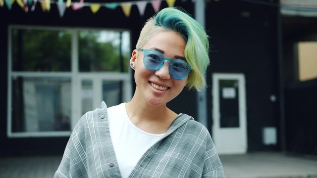 Asian young lady with dyed hair and nose piercing is waving hand and smiling standing outdoors alone. Welcome, greeting and friendly people concept.