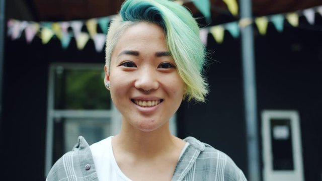 Close-up portrait of cute Asian hipster with dyed hair and nose piercing smiling outside looking at camera standing alone. Urban style and happy people concept.
