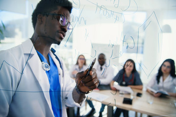 African professor at medical conference in modern clinic teaches his students. Doctor writing on board some formulas for interns in conference room at hospital at sunrise.