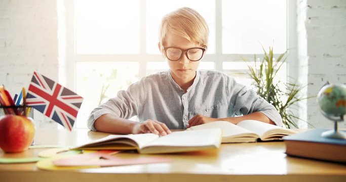 Smart boy in eyeglasses sitting in classroom and reading english textbooks in evening, preparing homework