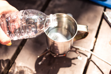 woman hand holding clear plastick bottle and pouring water in to the metal mug heating on gas-stove portable gas burner on a wooden table. Camping equipment