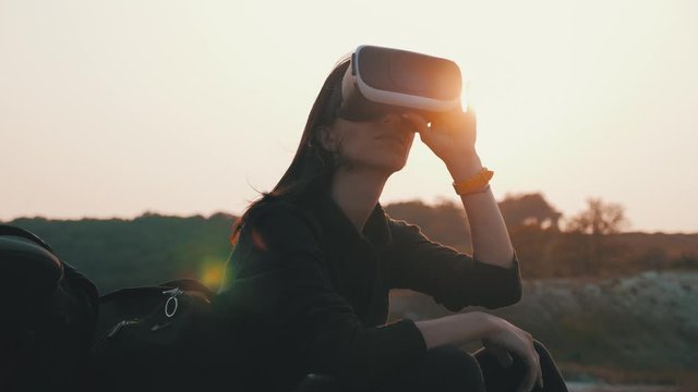 Hipster young girl with backpack on the top of a rock uses virtual reality glasses in the sunset. Active modern futuristic lifestyle leisure concept.