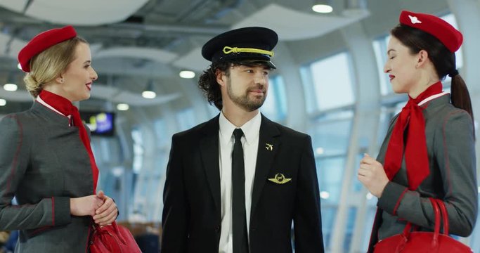 Handsome Caucasian pilot in a uniform and two beautiful young stewardessas talking cheerfully at the airport hall after arrival or before departure.