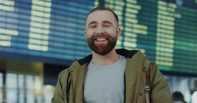 Portrait of the young handsome Caucasian guy with a berd standing in the airport and smiling to the camera on the flight timetable background. Close up.