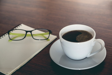 A cup of blackcoffee , blank notepad and eyeglasses on wooden table workpalce,