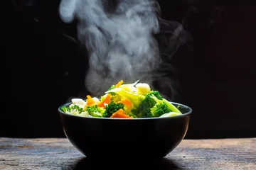 Fototapete Essen Der Dampf aus dem Gemüse Karotten Brokkoli Blumenkohl in einer schwarzen Schüssel, ein Dämpfen. Gekochtes heißes gesundes Essen auf dem Tisch auf schwarzem Hintergrund, warmes Essen und gesundes Essenskonzept