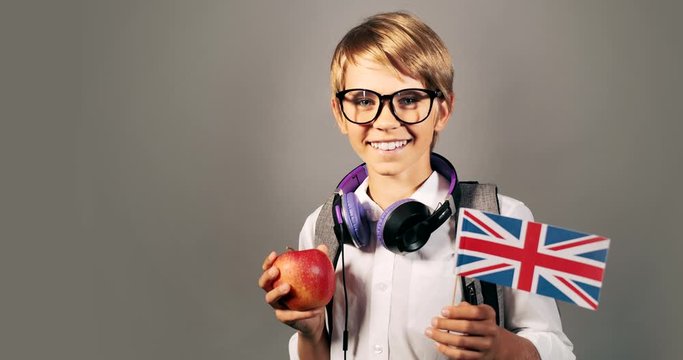 Smiling schoolboy in eyeglasses holding red apple and british flag looking to camera, isolated on grey background