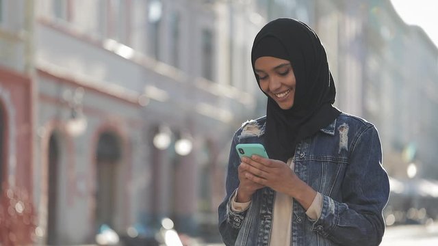 Young smiling muslim woman wearing hijab headscarf standing in the city center and using smartphone. Communication, online shopping, social network concept.