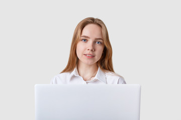 Fototapeta premium Photo of young Caucasian cheerful woman sitting over white wall with lportable aptop computer. Looking directly at camera with calm facial expression, working online, using wireless Internet.