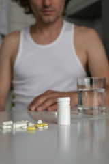 Pills and tablets and a glass of water on the table. A man sits in the kitchen. Sickness, malaise, headache, cold. Foreground. Vertical. Close-up