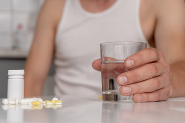 A man in home clothes sits at the white kitchen table, on which there are tablets, pills, and holds a glass of water. Illness, malaise, cold. Close-up