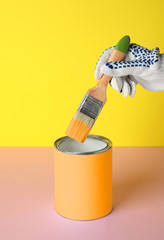 Person dipping brush into can of orange paint on pink table against yellow background, closeup....