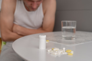 A man in home clothes sits on a chair and holds his hands on his stomach at the white kitchen table, on which there are tablets, pills and a glass of water. Severe abdominal pain, malaise. Close-up
