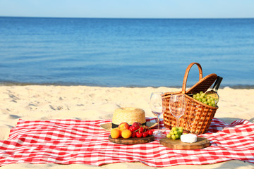 Checkered blanket with picnic basket and products on sunny beach. Space for text