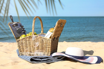 Wicker picnic basket with products, blanket and hat on sunny beach. Space for text