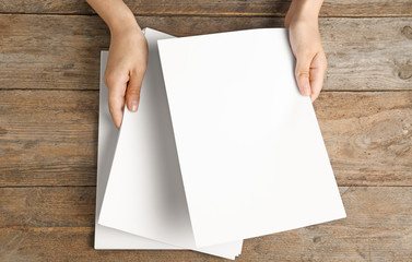Woman holding blank paper sheets for brochure at wooden table, top view. Mock up
