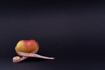 White-red measuring tape encircles a fresh ripe juicy apple on a black background, a tool for...