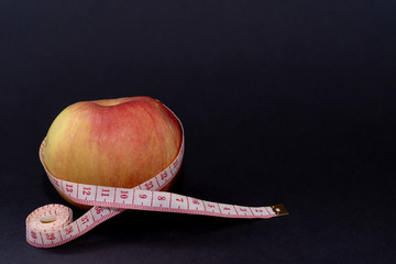 White-red measuring tape encircles a fresh ripe juicy apple on a black background, a tool for...