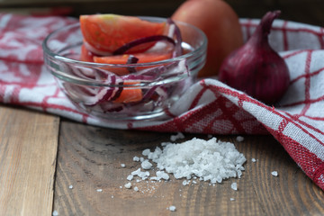 Close-up. A handful of sea salt, a salad of tomatoes and pink onion in a glass bowl with the addition of vegetable oil and a red and white napkin on a wooden table on a black background. 