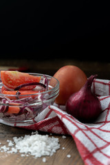 Tomatoes and pink onions, and a salad of them in a glass bowl with the addition of vegetable oil, a handful of sea salt and a red and white napkin on a wooden table on a black background. Vertical.