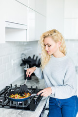 Fototapeta na wymiar Smiling attractive blonde woman frying vegetables in kitchen